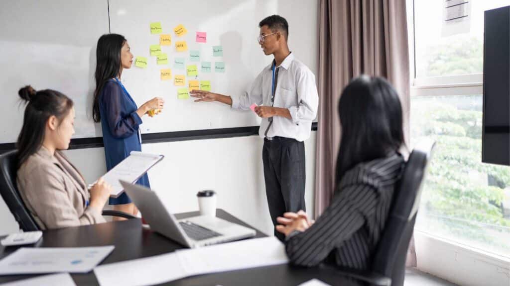 Modern office kitchen and dining area with white cabinets, wooden floors, and two dining tables with black chairs.