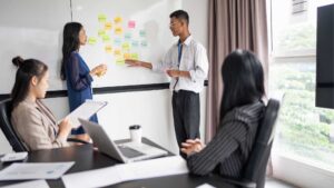 Modern office kitchen and dining area with white cabinets, wooden floors, and two dining tables with black chairs.