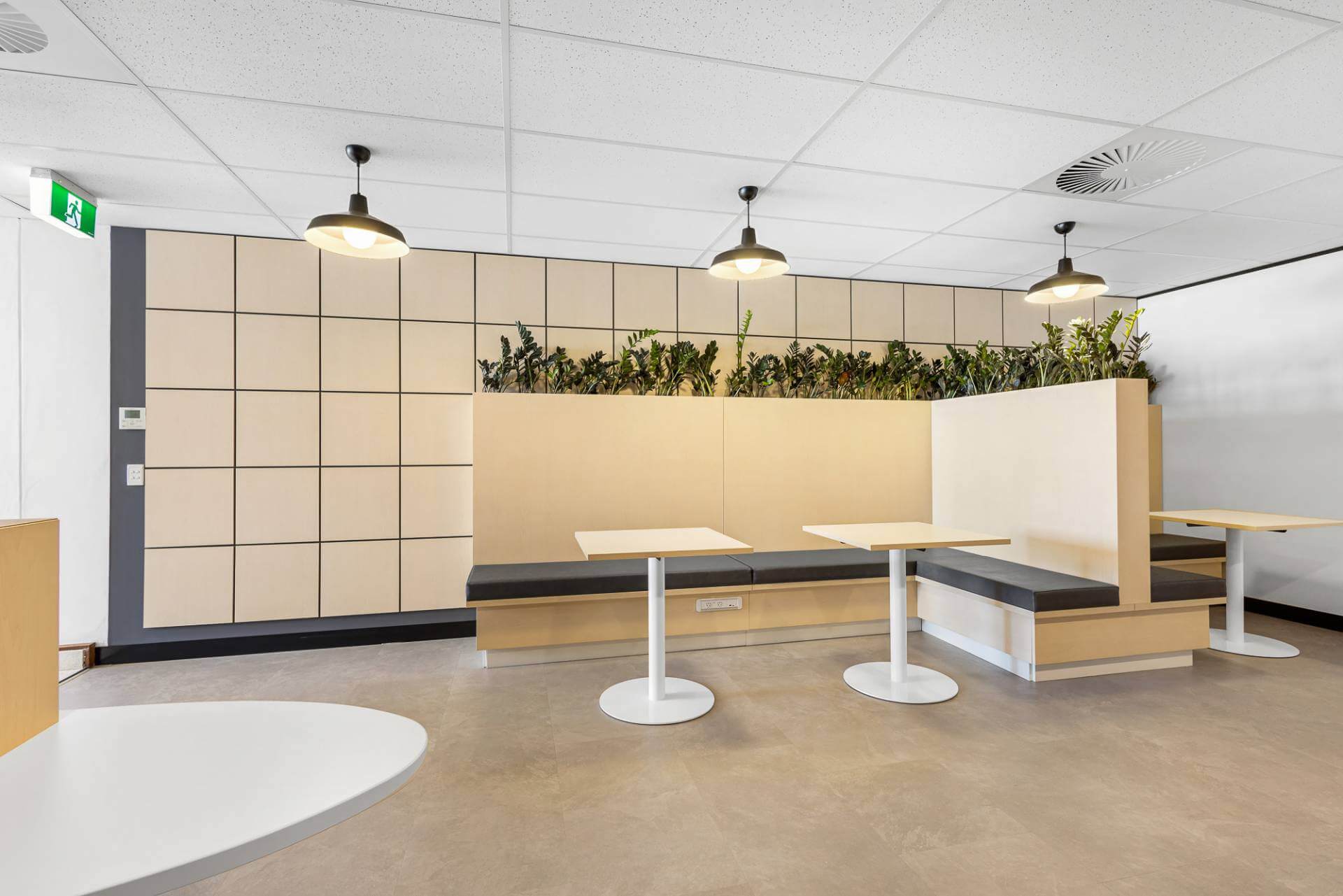 Modern office breakout area with light wood booths, white tables, and green indoor plants for a fresh, inviting atmosphere.