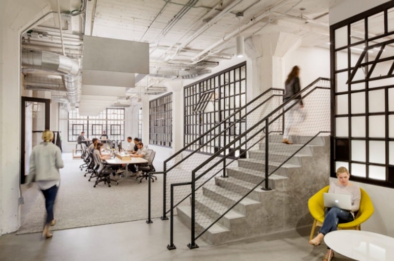 A high-ceiling, industrial-style office space featuring exposed ductwork, a concrete staircase with black metal railings, and large grid-style windows. Employees are seen working at a long communal table and in a cozy yellow lounge chair, reflecting a home-like, creative atmosphere.