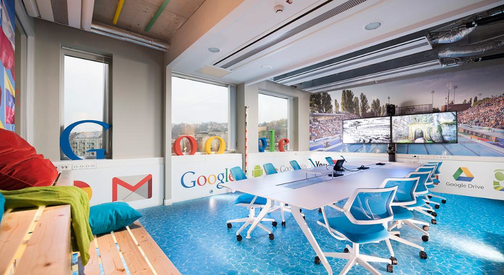 A vibrant Google conference room featuring a blue water-patterned floor, white modern chairs, and walls adorned with Google product logos like Gmail and Drive. Large windows offer a city view, and a large digital display sits at the head of the table.