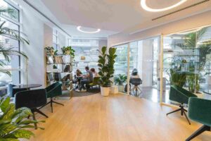 A brightly lit, modern office lounge featuring light wood flooring, glass partitions, and lush indoor plants. Employees sit in a collaborative breakout area with plush velvet green chairs and circular halo lighting, illustrating a high-end office fitout designed for employee well-being and success.
