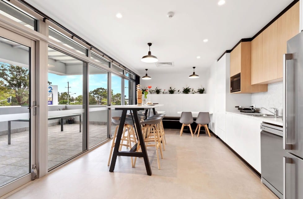 A clean, contemporary office kitchen and breakroom featuring white cabinetry, light wood accents, and a high communal table with grey stools. Large glass sliding doors lead to an outdoor terrace, encouraging spontaneous interactions and employee relaxation.